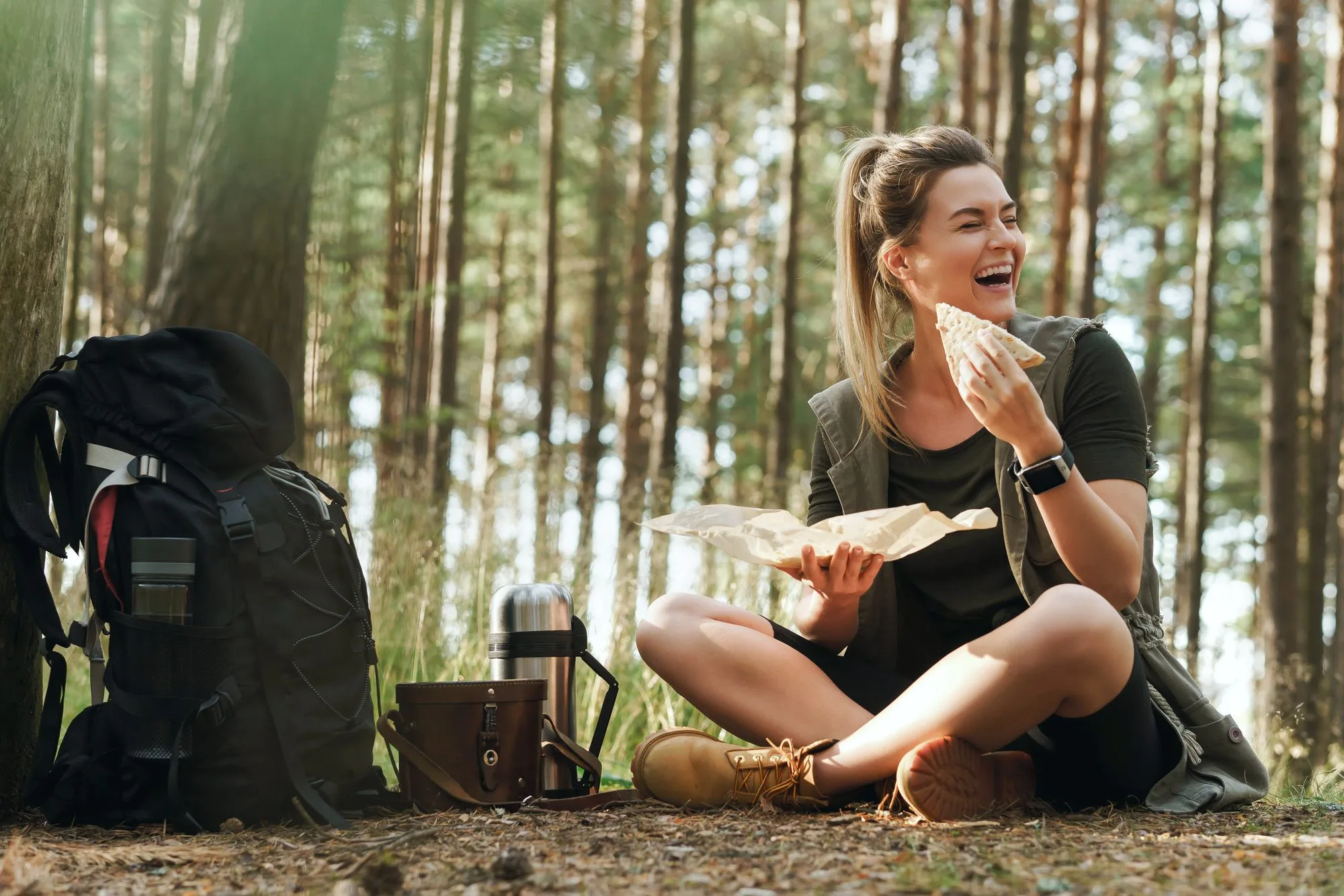 A walker laughing over lunch in a pinewood, rucksack and flask beside her